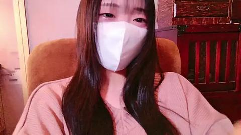 Media: A video of an East Asian woman with long black hair, wearing a white surgical mask, a beige sweater, and a light pink shirt, sitting in a mustard-colored armchair in a dimly lit room.