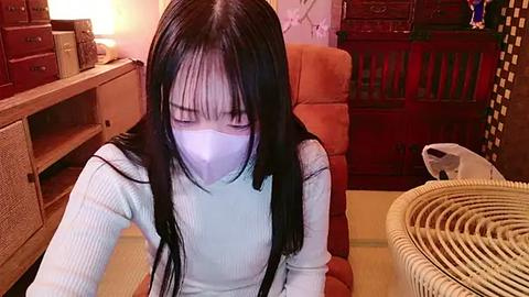 Media: Video of an Asian woman with long black hair, wearing a white mask, seated at a desk in a cozy, cluttered room with wooden furniture and a wicker basket.