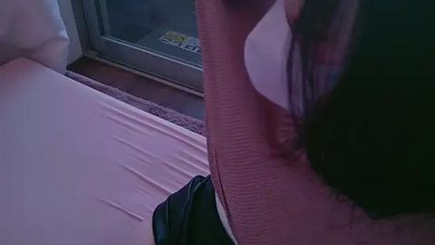 Media: A dimly lit video shows a woman with long black hair, wearing a white shirt, leaning against a window with a visible parking permit sticker. She sits on a bed with a light pink sheet.