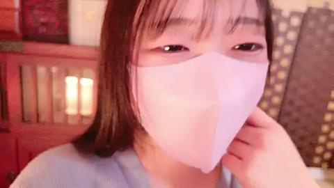 Media: Video of an East Asian woman with straight, shoulder-length brown hair, wearing a light blue shirt and a white surgical mask, sitting indoors with wooden furniture and patterned wallpaper in the background.