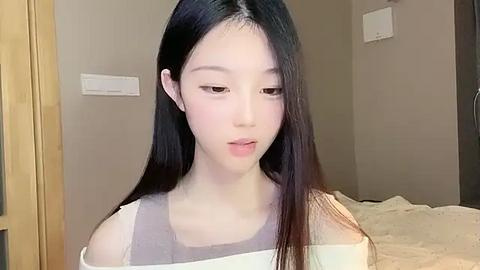 Media: A video of a young Asian woman with long black hair, fair skin, and a slender physique, wearing a white off-shoulder top, indoors against a beige wall.