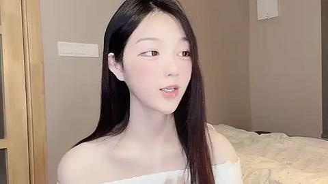 Media: Video of a young East Asian woman with long black hair, fair skin, and a slender physique, wearing a white off-shoulder top, standing indoors in a softly lit room with beige walls and a white bed.