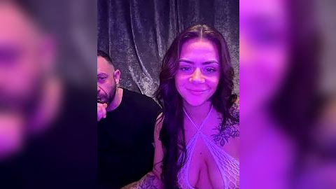 Media: Video of a smiling, tattooed woman in a revealing, crisscrossed top, standing next to a bearded man in a black shirt, both in front of a dark, textured curtain.