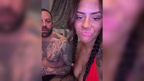 Media: A video shows a tattooed, shirtless man with a beard, and a woman with braided hair and a red top. They are indoors, with blurred background.