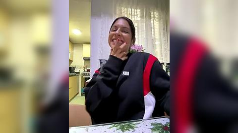 Media: A video of a smiling woman with dark hair, wearing a black jacket with red and white stripes, seated in a kitchen with light wood cabinets, white curtains, and a floral tablecloth.