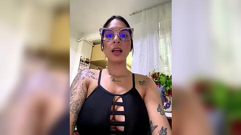 Media: Video of a tattooed, light-skinned woman with glasses, wearing a black halter top, indoors with a blurred background.