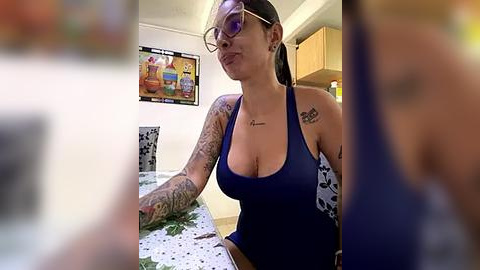 Media: A video of a tattooed woman with glasses, wearing a deep blue, low-cut tank top, sitting at a dining table in a kitchen.