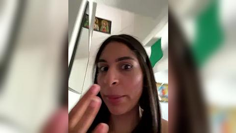 Media: A close-up video of a young woman with long dark hair, brown eyes, and light skin, making a \"V\" gesture with her fingers in front of a white refrigerator.