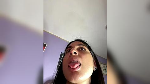 Media: A video of a young woman with long black hair sticking her tongue out, taken from a low angle, capturing her from below. The background shows a purple wall with a framed picture and a ceiling light.