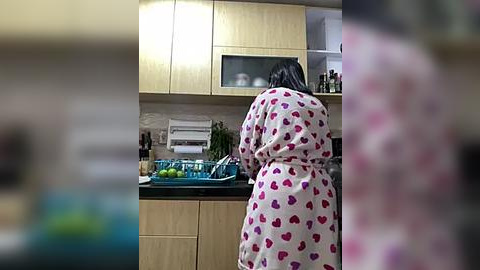 Media: Video of a woman with long black hair, wearing a white robe with red heart patterns, standing in a modern kitchen with light wooden cabinets, countertop cluttered with fruits, utensils, and green plants.