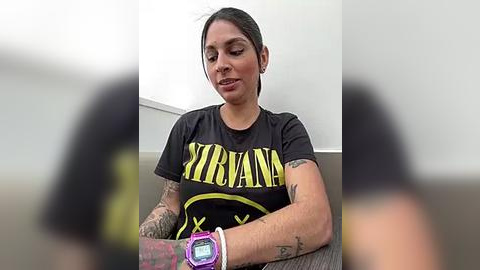 Media: Video of a tattooed woman with short brown hair wearing a Nirvana t-shirt and a purple smartwatch, sitting indoors. Background is blurred.