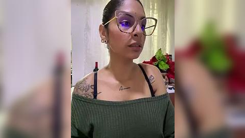 Media: A video of a light-skinned woman with a medium build, wearing glasses and an off-shoulder green top, with tattoos on her chest and arms, in a dimly lit room.