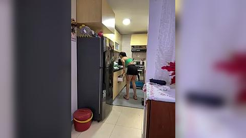 Media: Video of a woman in a black tank top and shorts cleaning a kitchen with white cabinets and stainless steel appliances. The background features a red stool and a white table with a floral cloth.