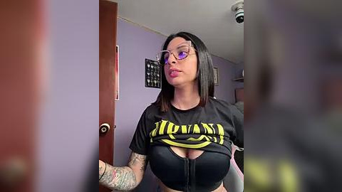 Media: Video of a tattooed woman with glasses and purple lipstick, wearing a black \"Hollywood\" crop top, standing in a purple-walled room.