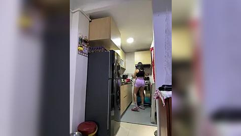 Media: Video of a small, cluttered kitchen with a person in pink shorts and a black tank top, standing in the middle, and a large black refrigerator on the left.