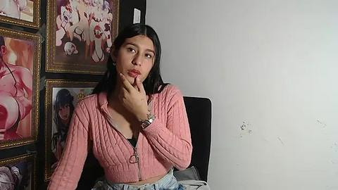 Media: Video of a young Latina woman with long black hair, wearing a pink zip-up top and high-waisted jeans, contemplating with her finger to her lips, in a room with colorful anime posters on the wall.