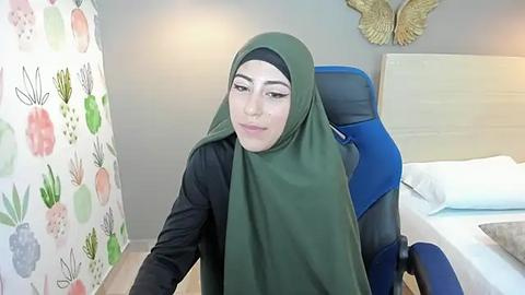 Media: Video of a Middle Eastern woman with light skin, wearing a black top and green hijab, seated in a blue office chair, in a minimalist room with a white bed and floral-patterned wall.