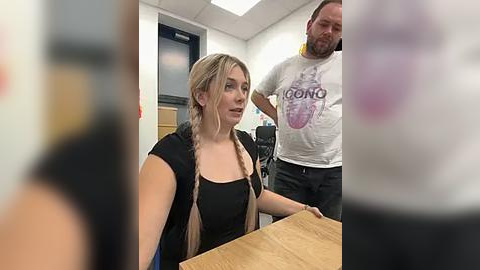 Media: Video of a blonde woman with braided hair in a black dress, sitting at a table, flanked by a bearded man in a white \"Goon\" t-shirt and another man in a black shirt. Background shows a classroom with a whiteboard and chairs.