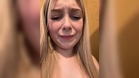 Media: A close-up video of a young Caucasian woman with long, straight blonde hair, fair skin, and closed eyes, possibly crying. She is topless. Background features a textured beige wall.