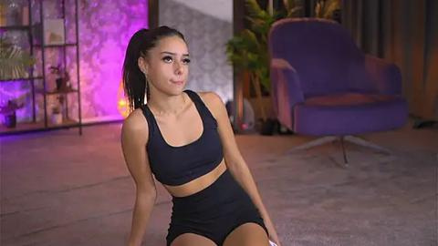 Media: Video of a fit woman with medium skin tone, long black hair in a high ponytail, wearing a black sports bra and shorts, sitting on a floor, indoors with a purple chair, bookshelves, and green plants in the background.