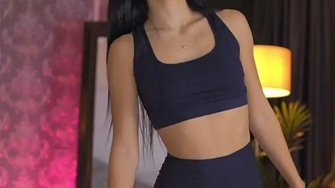 Media: Video of a fit, young woman with long black hair, wearing a dark blue sports bra and high-waisted leggings. Background features a warm-toned lamp, a pink wall, and a dark curtain.