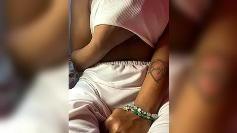 Media: Video of a woman with medium-brown skin, wearing a white ribbed crop top and loose white pants, sitting on a bed. She has a bracelet and a tattoo on her wrist. Background is blurred.