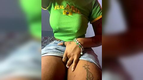 Media: A video of a woman with dark skin, wearing a green crop top and short denim shorts, revealing a tattoo on her inner thigh. Her hand is suggestively placed on her crotch.