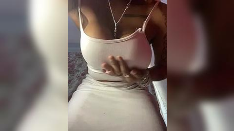 Media: Video of a woman with dark skin, wearing a tight beige dress, sitting on a bed, touching her chest.