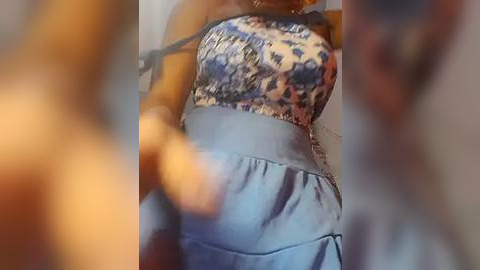 Media: Video of a woman with medium brown skin, wearing a blue and white patterned top and high-waisted blue skirt, taken from a slightly blurry angle.