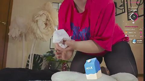 Media: A video shows a woman in a pink t-shirt and black leggings, pouring milk from a carton into a glass. Background features a plush lion, a plant, and a TV displaying video game elements.