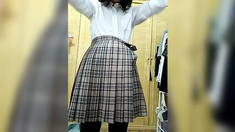 Media: Video of a person with medium-length dark hair, wearing a white button-up shirt and a pleated plaid skirt, standing in a bright, organized closet with wooden shelves and hanging clothes.