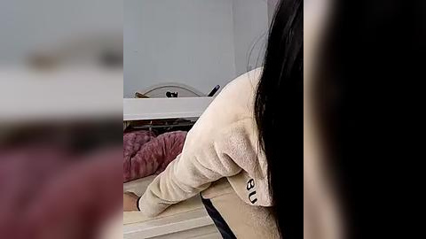 Media: Video of a person with long black hair wearing a beige sweatshirt, bending over a bed with a pink quilted blanket. Background features a white dresser and blurred objects.