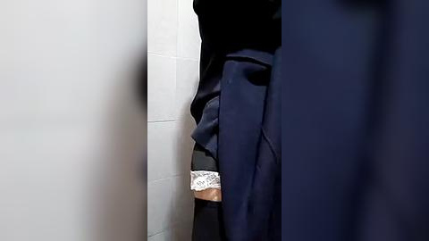 Media: A video of a person in a dark navy jacket, partially leaning against a white tiled wall, with a glimpse of a lace-trimmed white garment visible at the bottom.