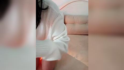 Media: Video of a woman in a white, long-sleeved sweater, sitting on a beige leather couch. Her long, dark hair cascades down her back. The background is softly lit, creating a cozy indoor atmosphere.