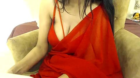 Media: Video of a light-skinned woman with long black hair, wearing a sheer, bright red halterneck dress, sitting in a beige armchair.