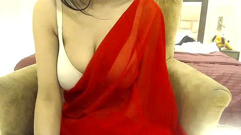 Media: Video of a woman with light skin, wearing a red saree draped over a white bra, sitting in a beige armchair in a modern bedroom with a bed and stuffed toy.