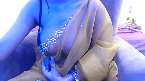 Media: A video of a light-skinned woman in a sheer, beige sari with intricate blue floral embroidery, partially exposing her left breast, sitting on a bed with pink sheets.