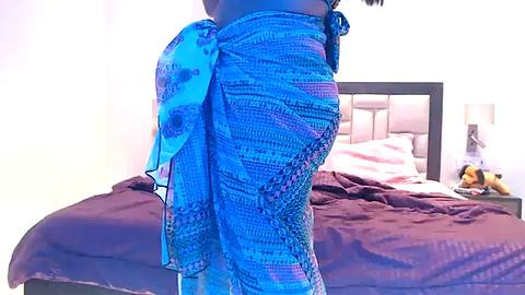 Media: Video of a person with dark skin wearing a blue patterned sarong, standing near a bed with maroon sheets and a beige headboard in a softly lit room.