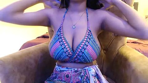 Media: Video of a woman with medium skin tone, wearing a vibrant, multicolored, patterned bikini top and matching skirt, seated on a brown, tufted chair in a softly lit, cozy room.