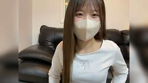 Media: Video of an Asian woman with long brown hair, wearing a white long-sleeve shirt with a subtle design, a white face mask, and sitting on a dark leather couch in a neutral-toned room.