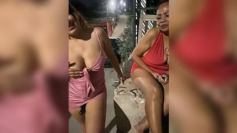 Media: Video of three people: two women and a man, one woman topless, all wearing pink clothing, outdoors at night, dim lighting, blurred background, handholding, intimate setting.