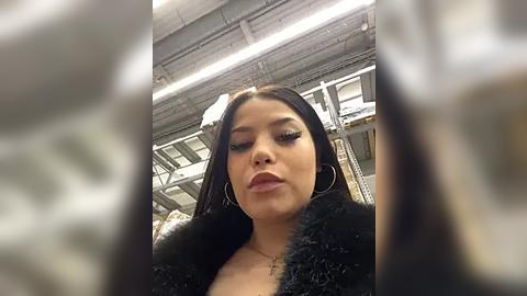 Media: Video of a Latina woman with long, straight black hair, wearing hoop earrings, a black fur coat, and bold red lipstick, looking serious. Industrial background with metal beams and fluorescent lights.