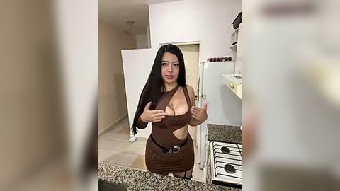 Media: Video of a Latina woman with long black hair, medium skin tone, wearing a brown, form-fitting dress that reveals her ample cleavage, standing in a modern kitchen with granite countertops.