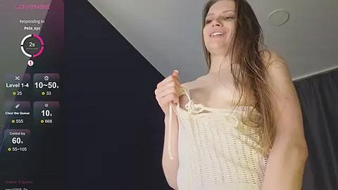 Media: A video of a wet, smiling woman with long brown hair, wearing a beige crochet top, standing in a dimly lit room with a dark wall and grey curtains.