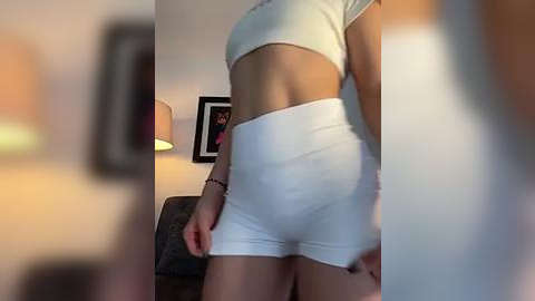 Media: Video of a woman in tight, white athletic shorts and a crop top, standing in a dimly-lit room with a framed picture on the wall and a lamp.