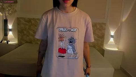 Media: A video of a woman wearing a \"Velvet Underground\" T-shirt, standing in a dimly lit, beige-tiled bedroom with two bedside lamps and a neatly made bed.