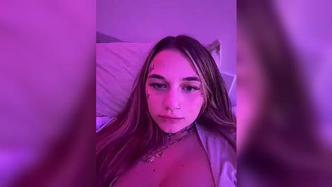 Media: Video of a young woman with long, straight brown hair, wearing a white robe, lying on a bed. She has fair skin, a nose ring, and a tattoo on her chest. The room is dimly lit with purple lighting, creating a moody ambiance.