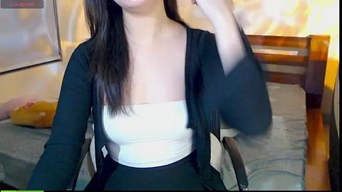 Media: Video of a young woman with long black hair, light skin, wearing a white tube top and black cardigan, sitting on a bed with a wooden headboard.