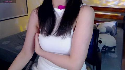 Media: Video of a young woman with straight, black hair, wearing a white tank top and pink choker, sitting on a black chair in a bedroom with a bed and stuffed animals.