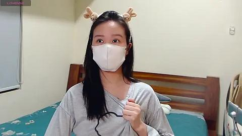 Media: Video of an Asian woman in a hospital bed, wearing a white mask, grey shirt, and bone-shaped headbands, giving a thumbs-up.